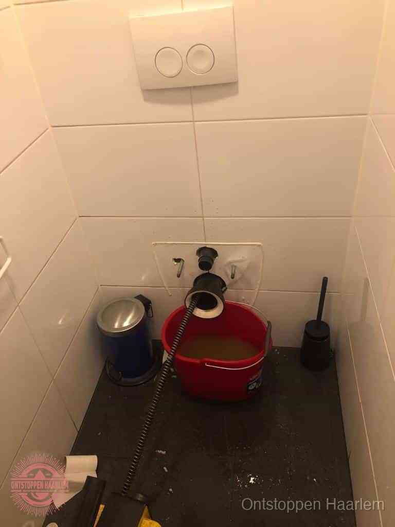 Een ontstopte wand-wc met verwijderde toiletpot waar een ontstoppingsveer in de wanduitgang verdwijnt, het achterhalen waar de verstopping zit in Haarlem