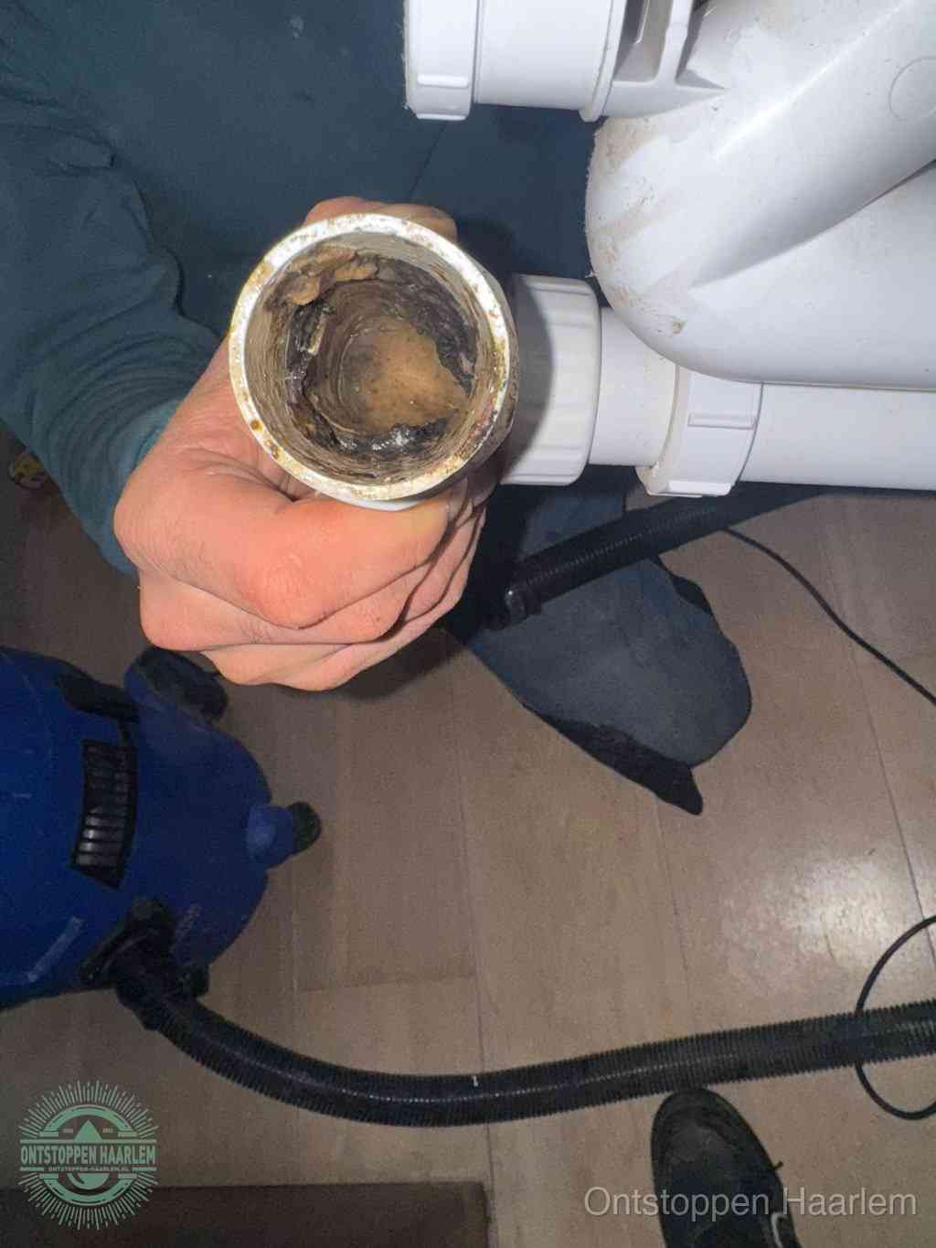 Een hand houdt een losgekoppeld PVC-afvoerbuisstuk vast dat van binnen volledig is dichtgeslibd met vet, vuil en aanslag, de meest voorkomende oorzaak van een verstopte afvoer in Haarlem